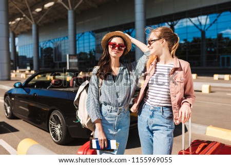 Young ladies in sunglasses holding passports with tickets and suitcase happily spending time together with cabriolet car on background