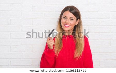 Young adult woman over white brick wall holding credit card with a happy face standing and smiling with a confident smile showing teeth
