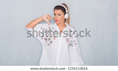 Young adult woman over grey grunge wall listening to music and dancing with angry face, negative sign showing dislike with thumbs down, rejection concept