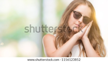 Young blonde woman wearing sunglasses sleeping tired dreaming and posing with hands together while smiling with closed eyes.