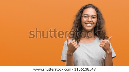 Beautiful young hispanic woman wearing glasses success sign doing positive gesture with hand, thumbs up smiling and happy. Looking at the camera with cheerful expression, winner gesture.