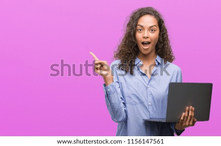 Young hispanic woman holding computer laptop very happy pointing with hand and finger to the side