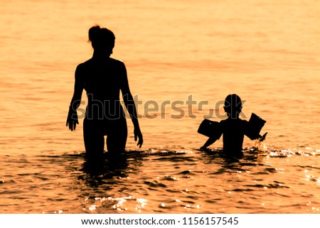 Silhouette of mother and son, the latter wears inflatable armrests, in the sea at sunset