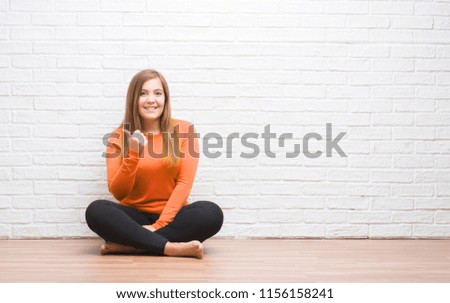 Young adult woman sitting on the floor in autumn over white brick wall happy with big smile doing ok sign, thumb up with fingers, excellent sign