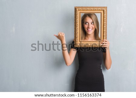 Beautiful young woman over grunge grey wall holding vintage frame pointing and showing with thumb up to the side with happy face smiling