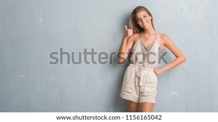 Beautiful young woman standing over grunge grey wall happy with big smile doing ok sign, thumb up with fingers, excellent sign
