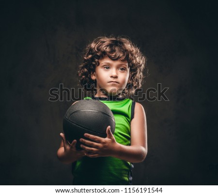 Little basketball player in sportswear holding ball in a studio. Isolated on the dark textured background.