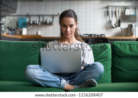 Serious woman using laptop checking email news online sitting on sofa, searching for friends in internet social networks or working on computer, writing blog or watching webinar, studying at home Royalty-Free Stock Photo #1156208407