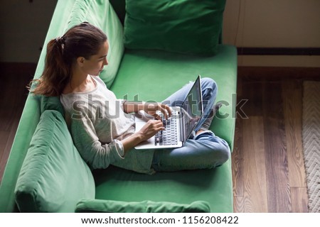 Relaxed woman using laptop sitting on sofa, millennial girl checking news in social networks, chatting with friends or working on computer, writing online blog or doing internet shopping at home Royalty-Free Stock Photo #1156208422