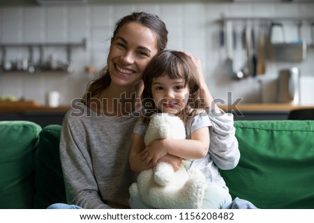 Portrait of happy family single mother and kid daughter embracing on sofa at home, young cheerful mom sister stroking cute girl hugging looking at camera, mum and child sincere relationship concept Royalty-Free Stock Photo #1156208482