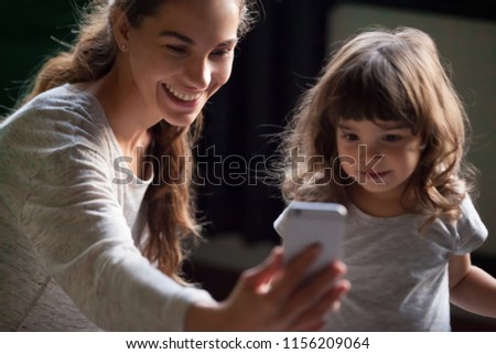 Smiling mother taking selfie on smartphone posing with cute kid daughter, happy young mom laughing making mobile photo with little girl at home, single mommy and child playing having fun with phone