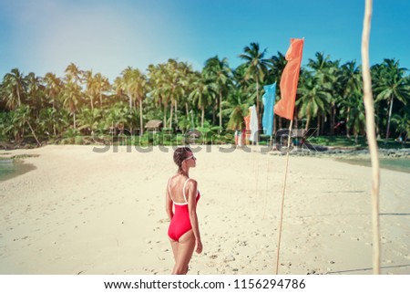 Vacation on the seashore. Back view of young woman in red swimsuit walking away on the beautiful tropical white sand beach.