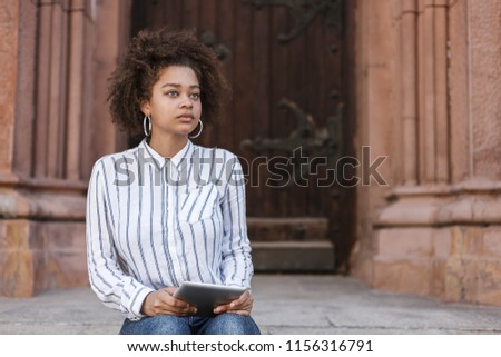 A dark-skinned girl sitting on the steps near the building. She looks straight and holds the tablet.