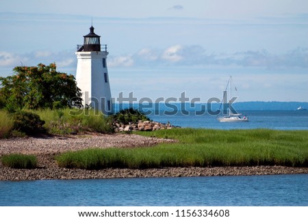 Sailboat passing by tower of Black Rock Harbor lighthouse, also referred to as Fayerweather Island light, near the rocky shoreline of Seaside Park, on a warm summer day in Bridgeport, Connecticut. Royalty-Free Stock Photo #1156334608