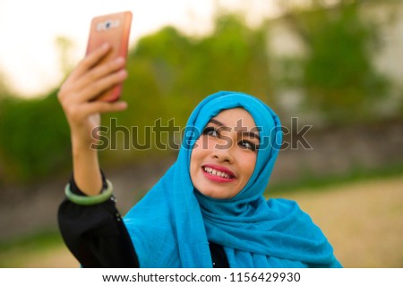 lifestyle portrait of young happy and beautiful tourist woman in muslim hijab head scarf taking selfie picture with mobile phone posing alone in holidays travel and smartphone technology concept