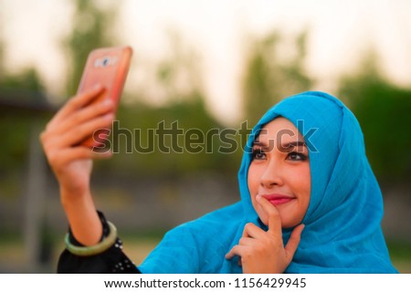 lifestyle portrait of young happy and beautiful tourist woman in muslim hijab head scarf taking selfie picture with mobile phone posing alone in holidays travel and smartphone technology concept
