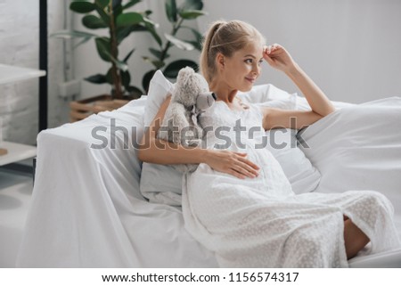 smiling pregnant woman in white nightie with teddy bear resting on sofa at home