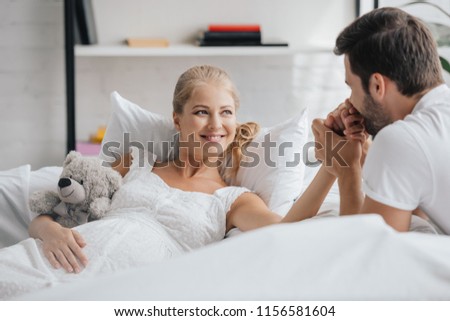 man and pregnant wife in white nightie on sofa holding hands at home