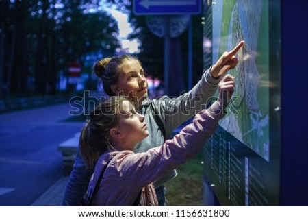 Two girls teenager are looking for the way route on the map scheme at night dark pointing their fingers to the scheme plan map