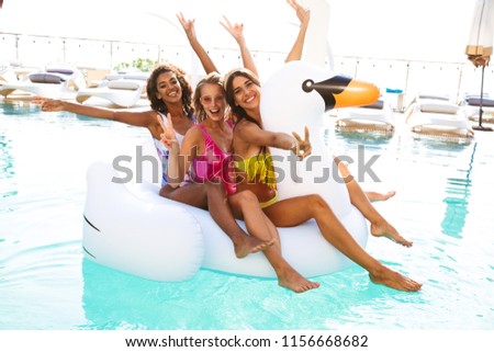 Three cheerful young women in swimwear swimming together at the swimming pool outdoors with inflatable ring, having fun, arms raised