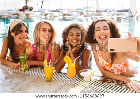 Four cheerful young women in swimwear taking a selfie while drinking cocktails together at the swimming pool outdoors