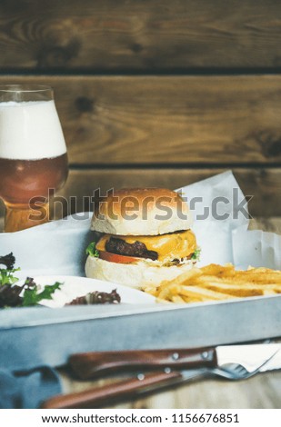 Classic burger dinner. Beef meat homemade burger with glass of lager beer, French fries and salad on tray, rustic wooden wall at background, copy space