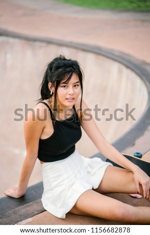 A young Chinese Asian skater girl (teenager, millennial) sits next to her skateboard in a skate park during the day and is listening to music on her smartphone with her bluetooth headset.