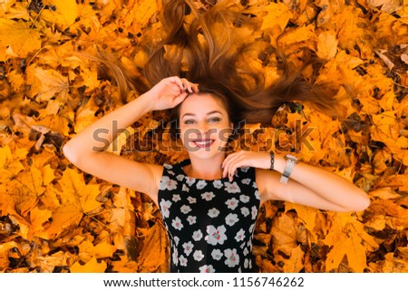 Beautiful and cute girl with long hair in autumn park on the sunny day lying in a big pile of leaves, top view
