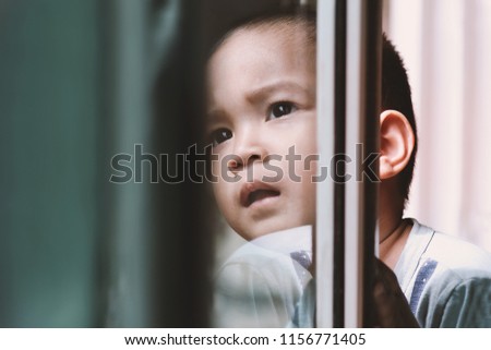 2 year old Asian toddler boy with skinhead hair style standing at the door and pokes his head through the gap of the sliding door.Kid during Covid-19 locked down or self isolated,Self quarantine.