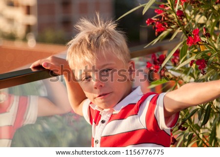 blond boy looks at the camera and throws his hand up with a victory gesture standing on the balcony of a hotel near the sea at sunset
