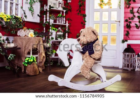 teddy bear rides a rocking horse in the yard of a  red wooden house