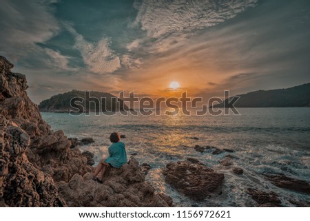 A girl sitting on the rocks looking at sunset, at beach, at sea