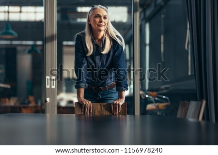 Portrait of successful businesswoman standing leaning to a chair in meeting room and looking away. Senior female entrepreneur in conference room. Royalty-Free Stock Photo #1156978240