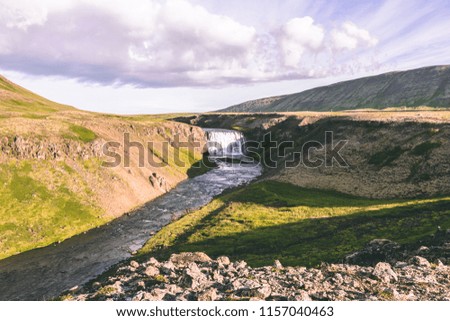 Waterfall in Iceland, river between cliffs, green grass lava fields, mountains and blue sky with clouds on the background