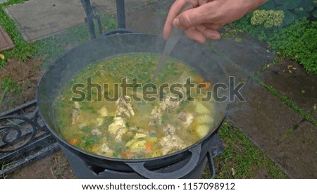 Delicious chicken soup is cooked on an open fire in a black cast-iron pot. A man's hand tries a soup with a ladle. Street food.
