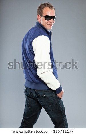 Happy young man with short hair wearing blue baseball jacket and blue jeans. Wearing black sunglasses. Sportive casual look. Studio shot isolated on grey background.