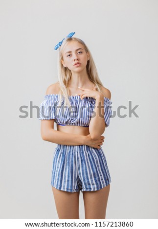 Calm and thoughtful beautiful blonde girl in summer outfit is standing, over white background. Emotions, gestures and facial expression