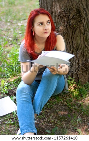 Student girl checking her diploma project while sitting with a mobile phone at university park. Travel blogger reading information in papers and checking it online while relaxing in a city park.