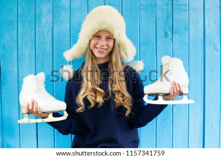 Cheerful little girl in warm sweater and hat keeps figure skates. Blue wooden background. 