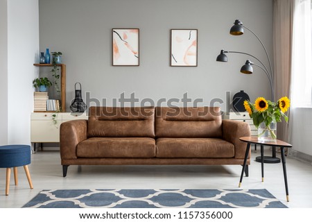 Sunflowers on wooden table next to leather sofa in living room interior with posters. Real photo Royalty-Free Stock Photo #1157356000