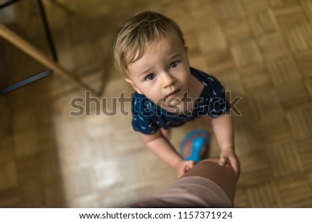 First steps of small baby. Portrait of a boy holding on mum's leg while standing at home. Blonde ten month age baby with blue body between mother legs looking at camera.