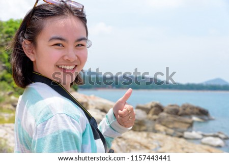 Young woman raised thumb up was happy to photography with dslr camera on the rock near the sea under the summer sky at Koh Samui island, Surat Thani province, Thailand