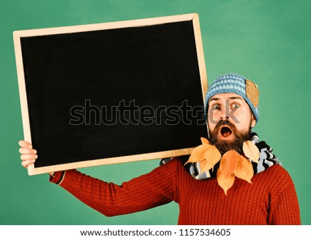 Autumn season sale concept. Guy holds chalkboard at barbershop, copy space. Man in warm hat holds blackboard on green background. Hipster with shocked face and beard full of autumn leaves