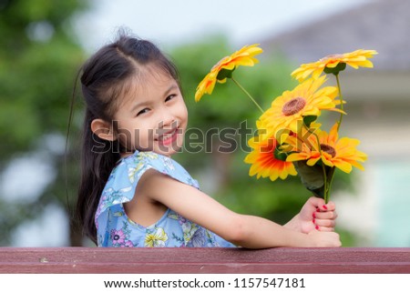 Young beautiful little asian girl holding and smelling sunflower in playground. Young girl with flower in blue dress smiling and happiness. Portrait cute cheerful young kid in the garden concept.
