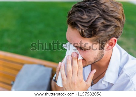 Man with allergy or an infection sneezing. Handsome hipster blowing his nose on a sunny day.  Flu season, vaccination. Isolated outside green trees background