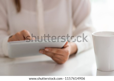 Businesswoman checking out financial news and drinking morning coffee. Closeup of woman sitting at white desk and using tablet near white mug. Business morning concept