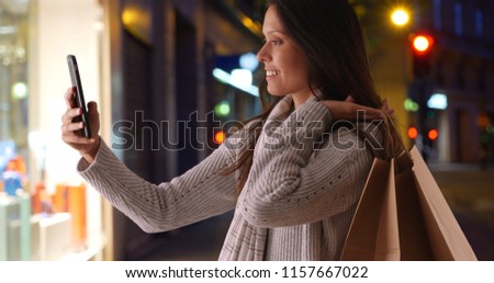 Millennial woman with shopping bags takes phone selfie on city street at night
