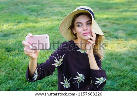  Young beautiful woman in trendy hat taking selfie on her phone on a green grass in a park