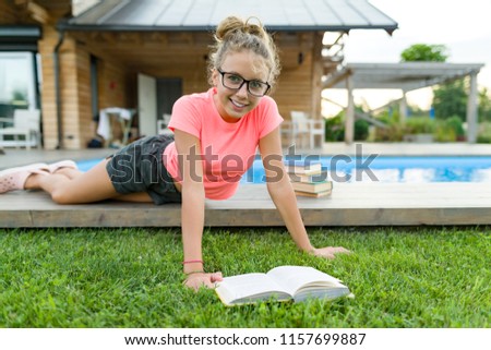 Teenage girl in glasses reads a book, background swimming pool, lawn near the house. School, education, knowledge, adolescents