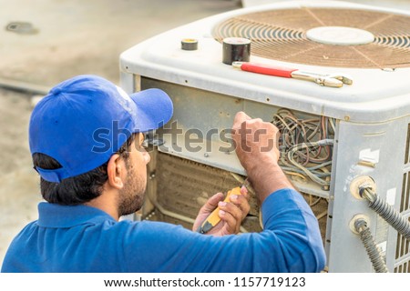 a professional electrician man is fixing the heavy air conditioner by his tools on the roof top and wearing blue color of uniform and head cap